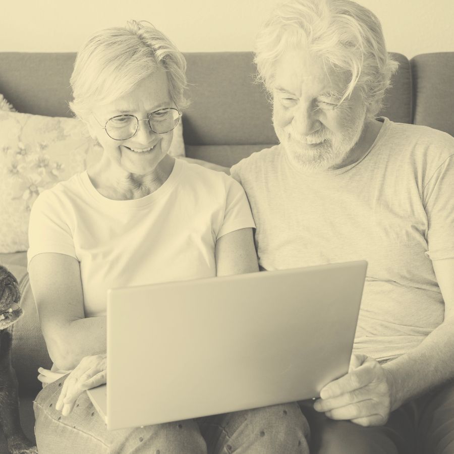 Ein älteres Paar sitzt auf einem Sofa und schaut gemeinsam auf einen Laptop. Die Frau trägt eine Brille und lächelt, während sie auf den Bildschirm zeigt. Der Mann, ebenfalls mit Brille, scheint konzentriert zuzuschauen.