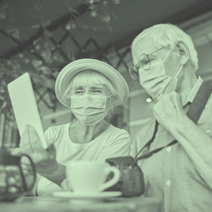Zwei ältere Menschen, beide mit Gesichtsmasken und in einem Café sitzend, schauen auf ein Tablet. Die Frau trägt einen Hut und scheint das Tablet zu bedienen, während der Mann zuschaut.