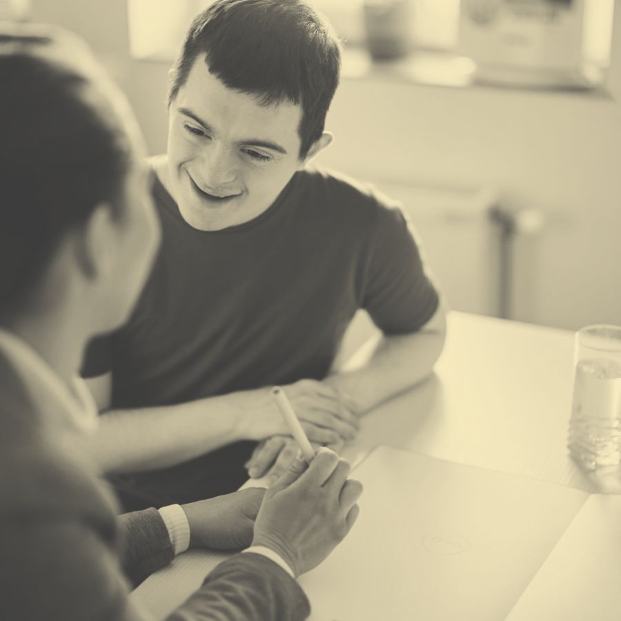 Ein junger, fröhlicher Mann mit Down-Syndrom sitzt an einem Tisch und unterhält sich mit einer ihm zugewandten Person, die lächelt und seine Hand hält.